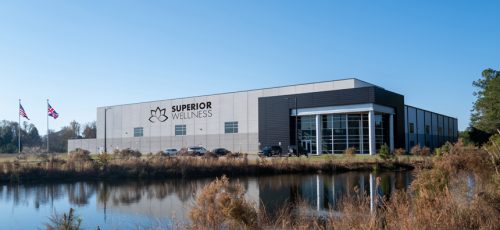 Exterior view of the Superior Wellness headquarters, a modern industrial building with large windows, situated near a pond with flags and natural landscaping in the foreground.