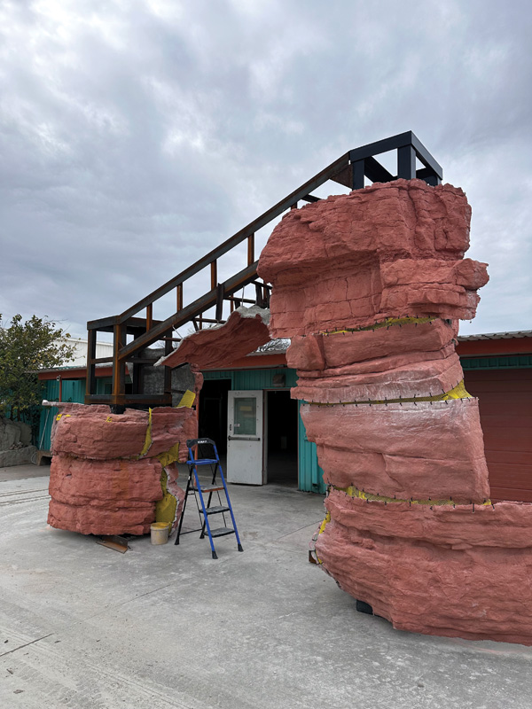 A large faux rock archway with a metal framework stands outside a building, with a ladder and tools nearby, under a cloudy sky.