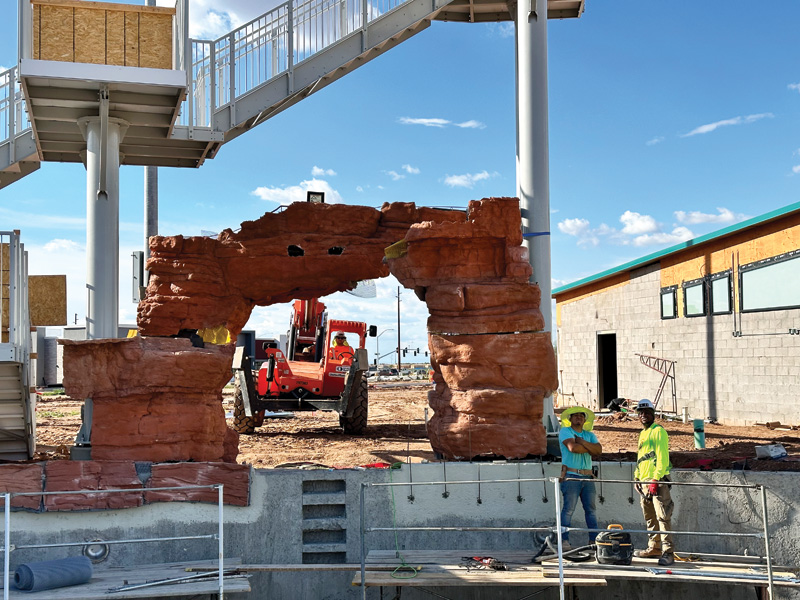 Two construction workers in safety gear stand near a large faux rock arch, with a lift and building materials in the background.