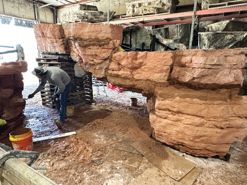 A worker applies finishing touches to a large, rocky structure crafted from simulated stone in a workshop setting.