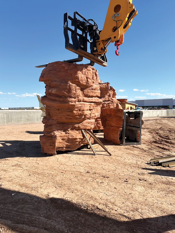 A construction site features a large, reddish rock formation being lifted by a yellow excavator in a clear blue sky.