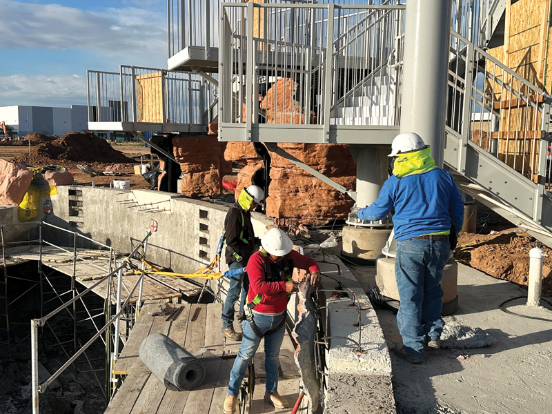 Three construction workers on a site, wearing hard hats, are overseeing structural work on a concrete platform with scaffolding.