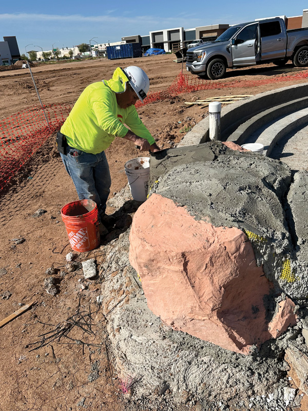 A construction worker in a neon yellow shirt applies concrete to a large rock structure at a construction site with vehicles in the background.