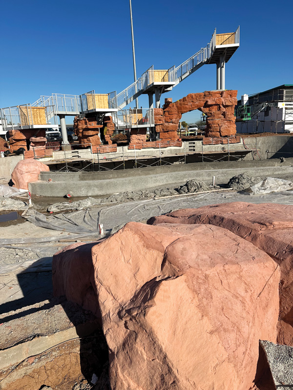 Construction site featuring pink rock formations, a view of an elevated walkway, and a partially completed concrete water area.