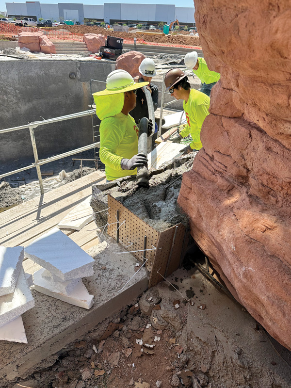 Construction workers in bright yellow shirts are pouring concrete into forms on a job site surrounded by rocks and building materials.