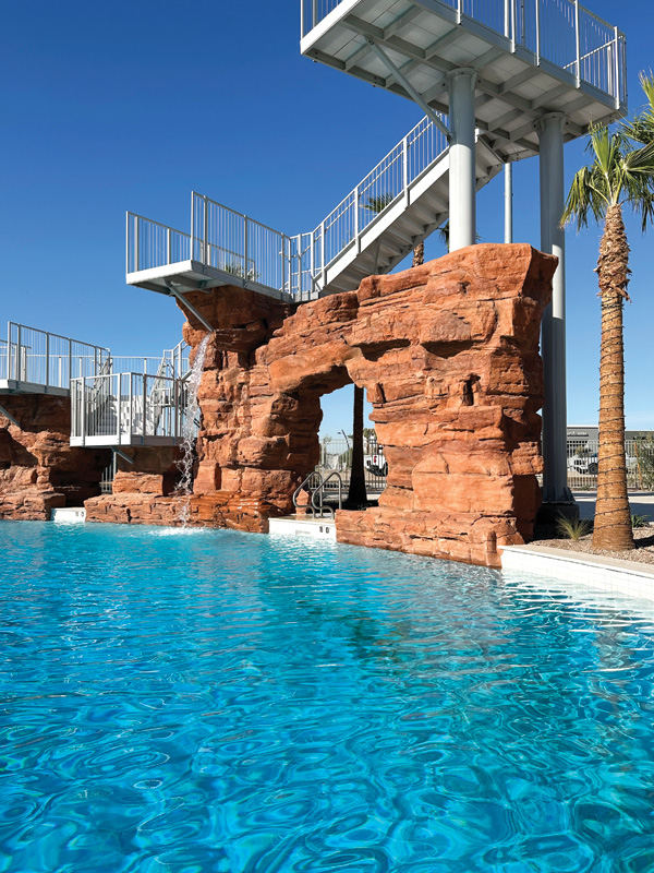 A vibrant swimming pool features a rock formation and a water slide, surrounded by palm trees under a clear blue sky.