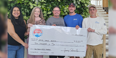 A group holds a large check for $10,000 made out to Child Crisis Arizona, surrounded by lush greenery indoors.