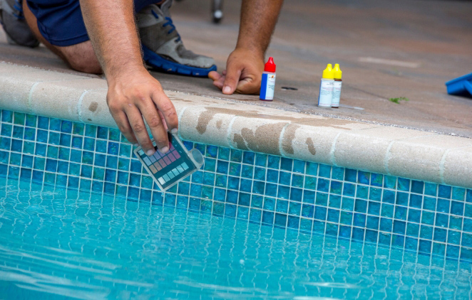 A person testing pool water with a colorimetric strip near the water's edge, with chemical bottles visible nearby.