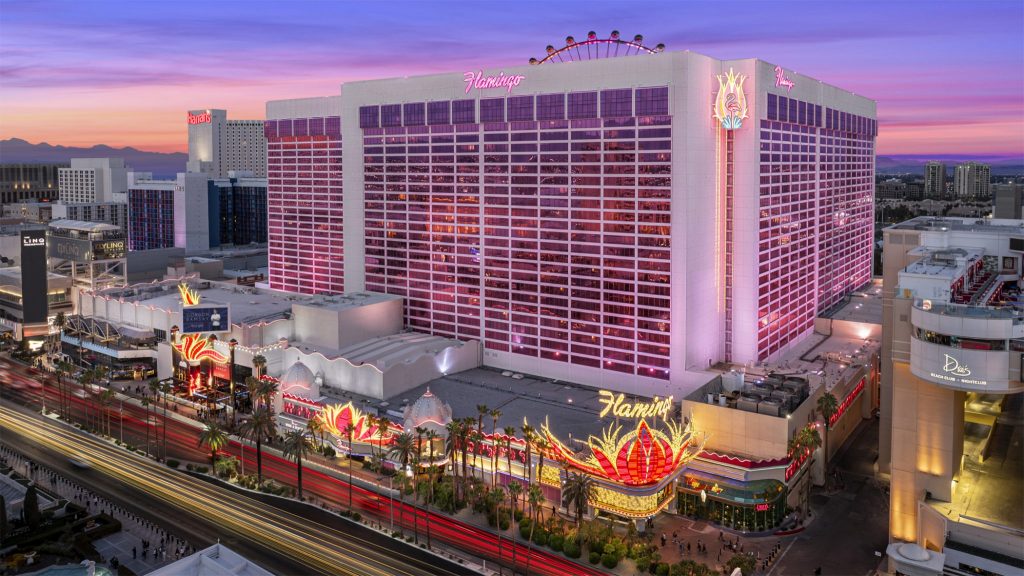 Aerial view of the brightly lit Flamingo Las Vegas hotel, showcasing its colorful facade and surrounding palm trees at sunset.