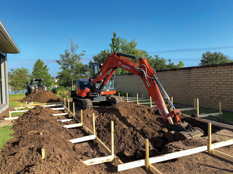 Two excavators work on a dirt-filled construction site, with wooden frames and a clear blue sky in the background.
