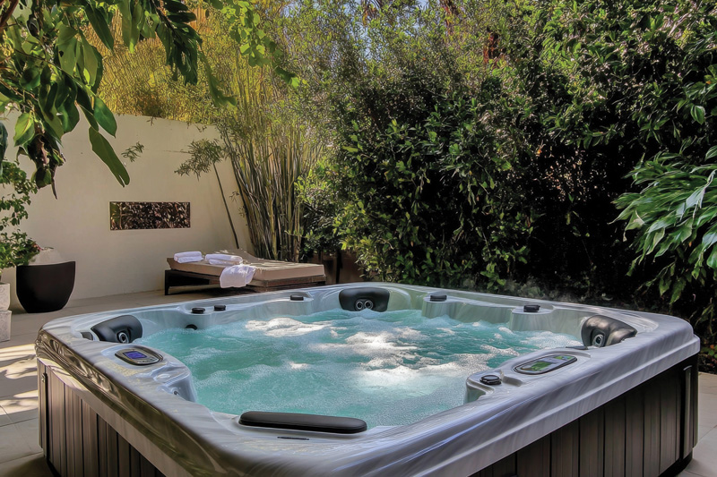 A bubbling hot tub surrounded by lush greenery and a sunlit lounge chair with rolled towels, creating a tranquil outdoor retreat.