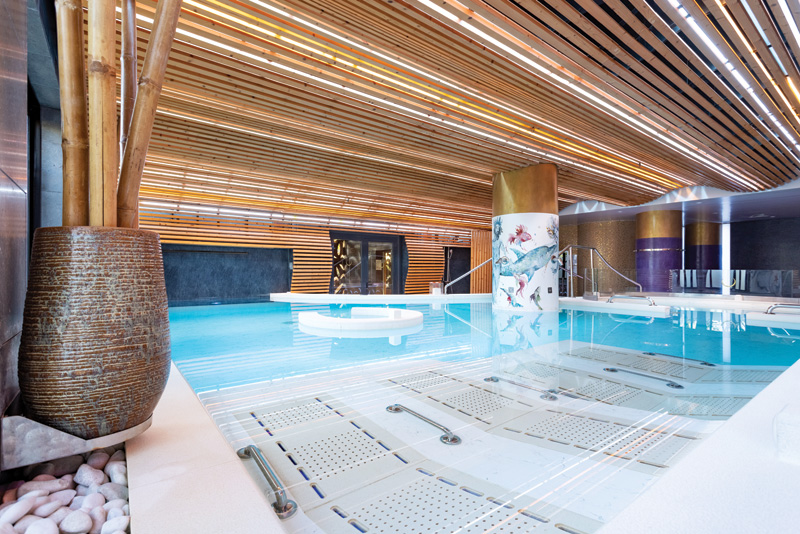 Modern indoor pool with wooden slat ceiling, adorned with bamboo and artistic column, creating a serene, luxurious atmosphere.