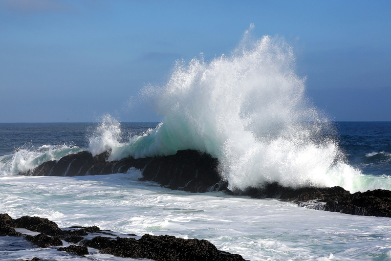 A powerful ocean wave crashes against rocky shoreline, sending up a spray of white foam under a clear blue sky.