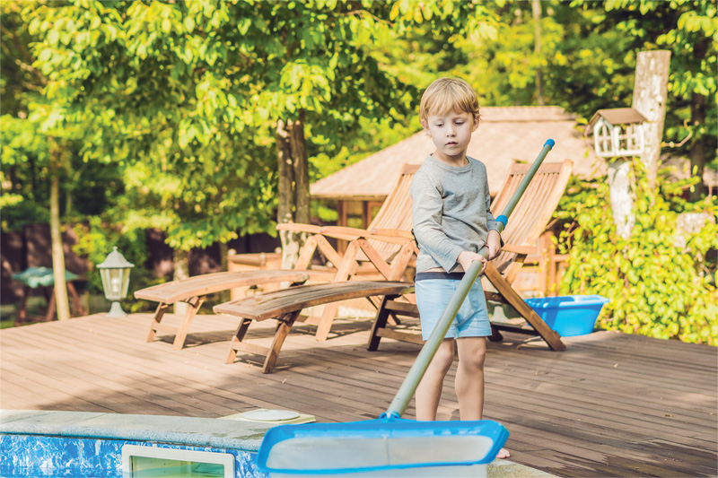 A child stands on a wooden deck near a pool, holding a skimmer net, surrounded by green trees and patio furniture in a sunny setting.