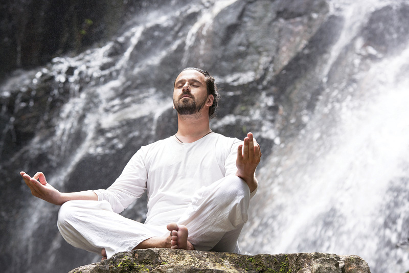 A person in white clothing meditates peacefully on a rock beside a cascading waterfall, surrounded by lush greenery.