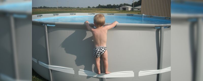 A toddler in checkered swim trunks stands on the edge of an inflatable pool, reaching over the water, with a sunny landscape behind.