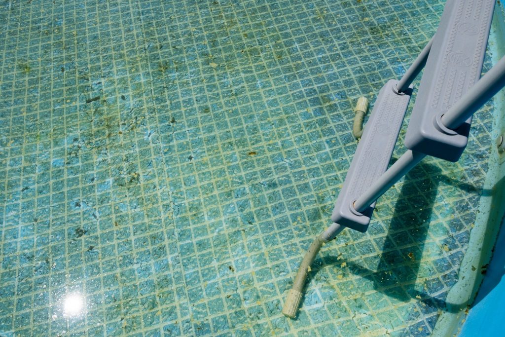 A partially submerged gray ladder in a turquoise pool with visible debris and a sunlit reflection on the water's surface.