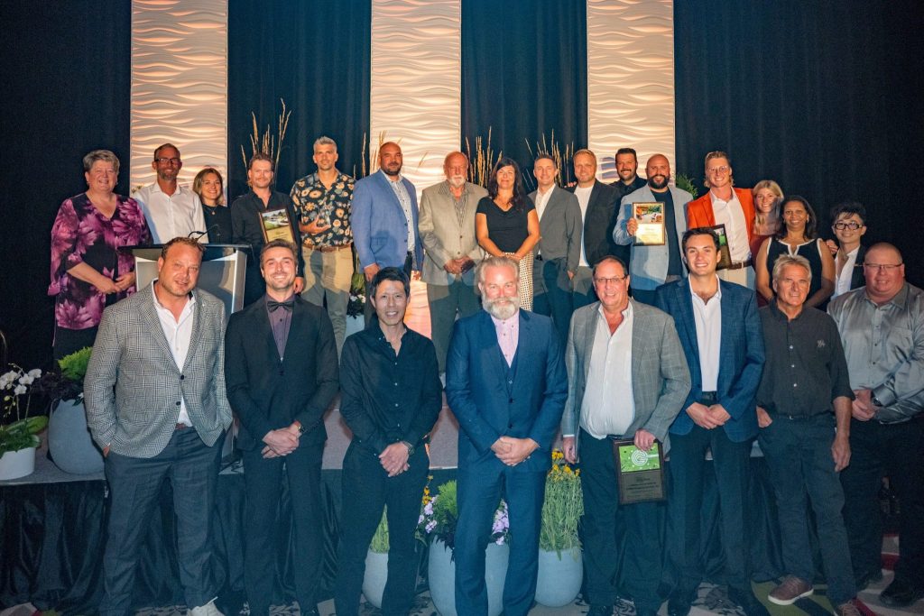 Group photo of attendees at an event, posing on stage with awards, surrounded by decorative plants and a stylish backdrop.