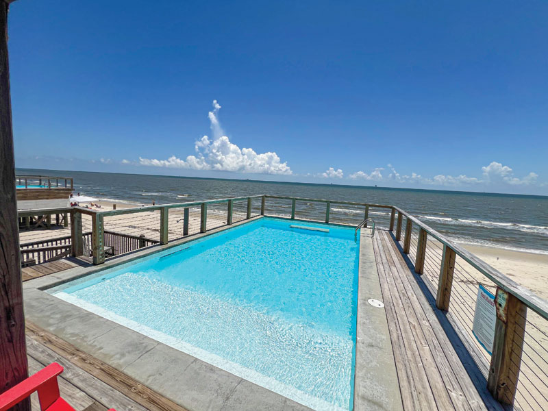 A sunny beachside view featuring a bright blue pool with wooden deck overlooking the ocean and distant clouds in a clear sky.