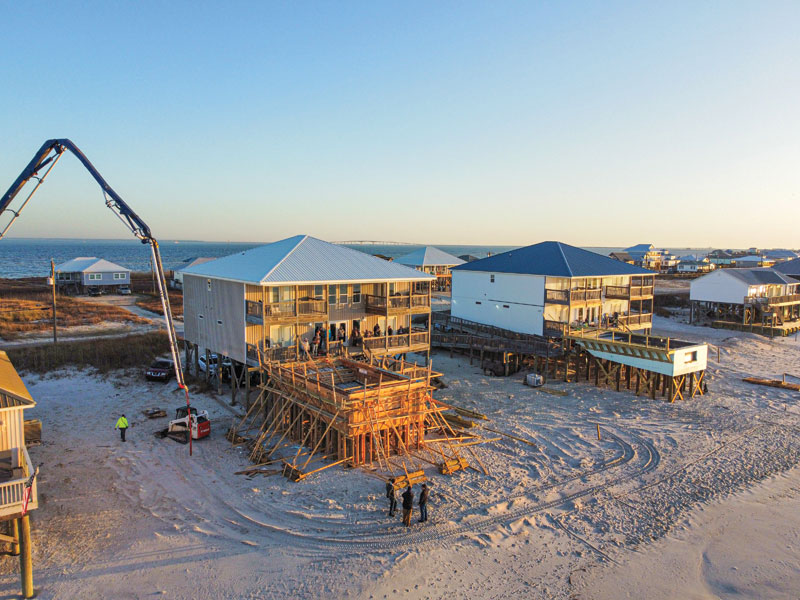 Construction workers on a beach site strand, operating a concrete pump for elevated homes at sunset, surrounded by sandy terrain.