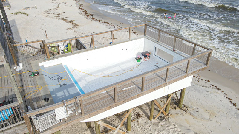 Aerial view of a beachside pool under construction, with workers inside and ocean waves nearby, surrounded by a wooden deck.