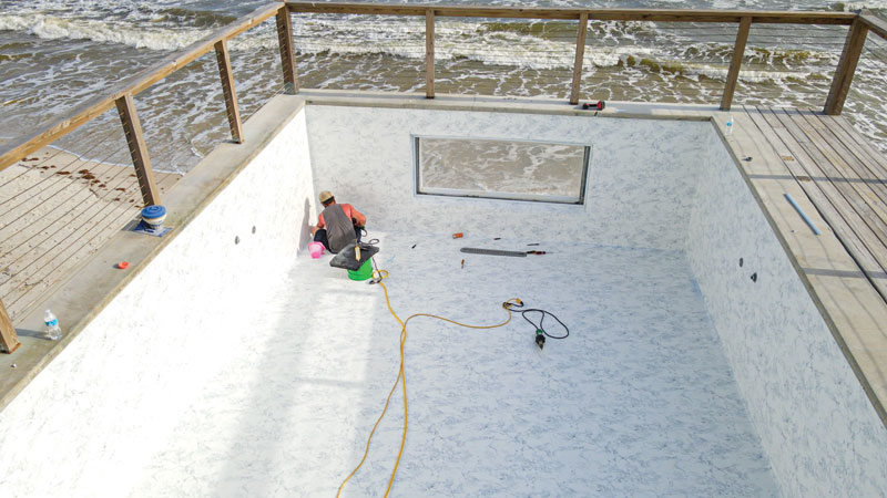 A worker installs a white marble-patterned liner in a swimming pool overlooking the ocean, with tools scattered around.