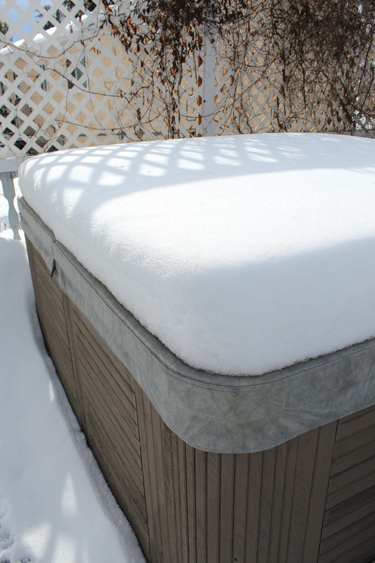 A hot tub covered with a thick layer of snow, surrounded by a white lattice fence and shadow patterns from nearby plants.