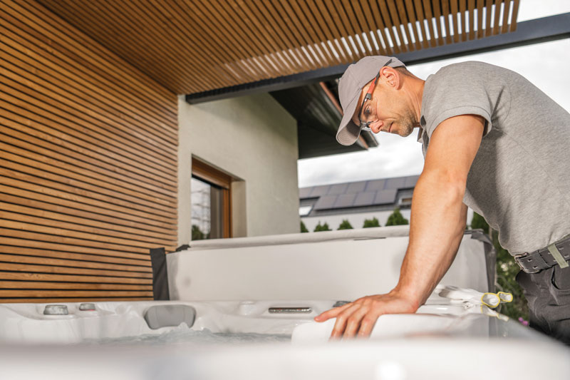 A person leans over a hot tub, inspecting its water, surrounded by a modern outdoor setting with wooden accents and a cloudy sky.