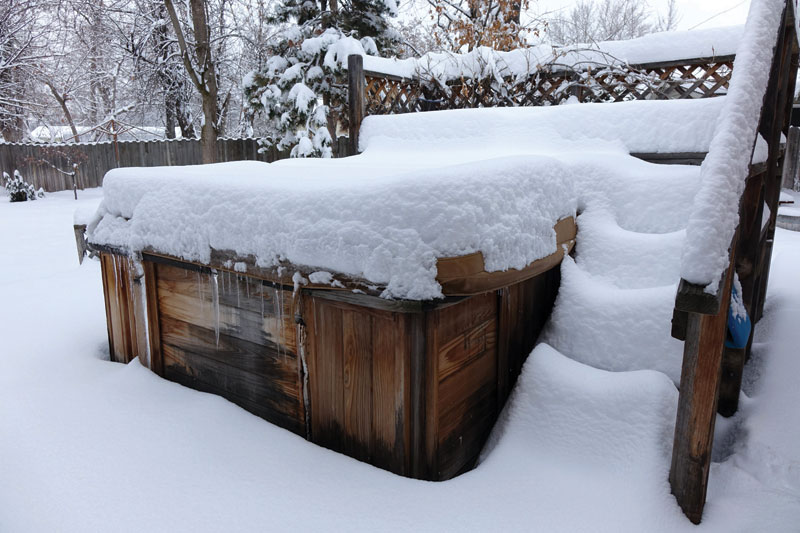 A hot tub covered in heavy snow, with snowdrifts surrounding the steps and deck, under a cloudy winter sky.