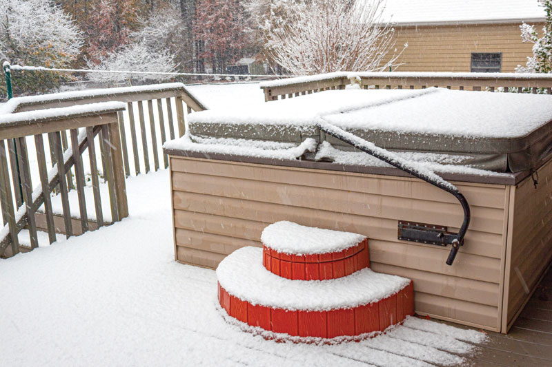 A hot tub covered in snow with red steps, set on a deck amidst a winter landscape of falling snowflakes and frosted trees.