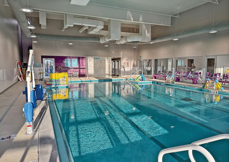 Indoor swimming pool with clear water, pool cleaning equipment, purple benches, and large windows letting in sunlight.