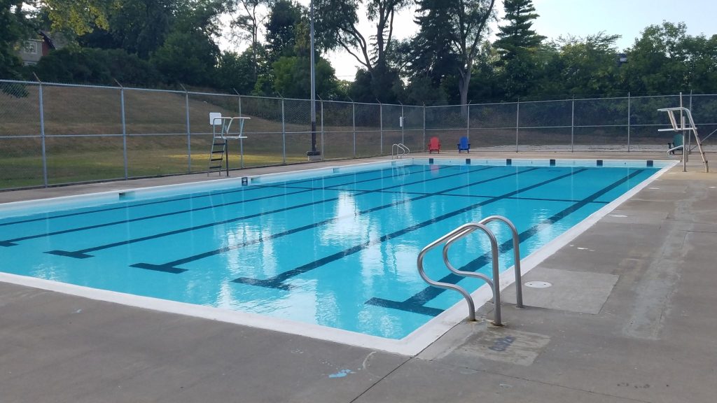 Outdoor swimming pool with clear blue water, surrounded by a concrete deck and chain-link fence. Ladders and lifeguard chairs are visible. Calm and serene setting.
