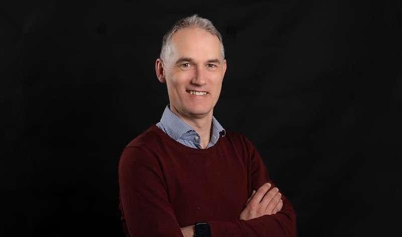 A smiling man with short gray hair stands against a black background, wearing a red sweater over a blue shirt, arms confidently crossed.