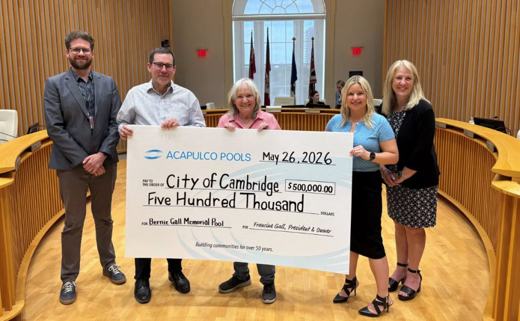 A group of five people stands in a formal room holding a large check for $500,000, dated May 26, 2026, from Acapulco Pools to the City of Cambridge.