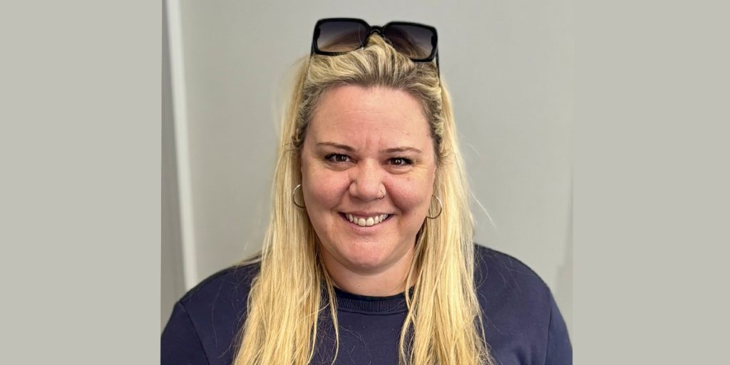 Smiling woman with long blonde hair wearing a navy top and sunglasses on her head against a plain background. Friendly and approachable expression.
