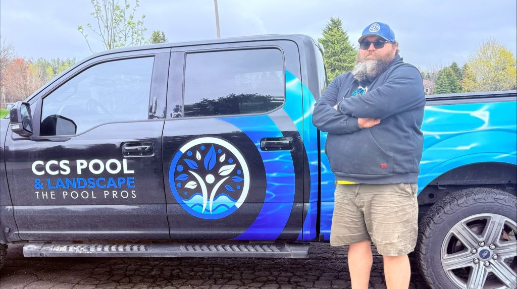 A man stands confidently with crossed arms beside a black and blue pickup truck. The truck features a logo and text reading 
