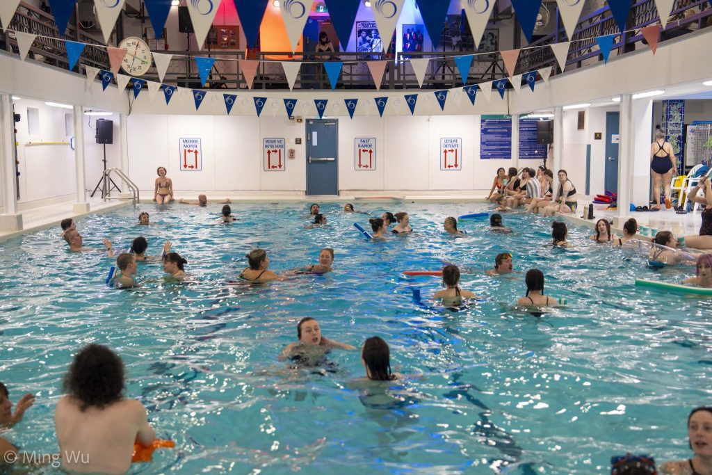 A busy indoor swimming pool with people enjoying the water. Colorful flags hang above, and a lively, social atmosphere fills the scene.