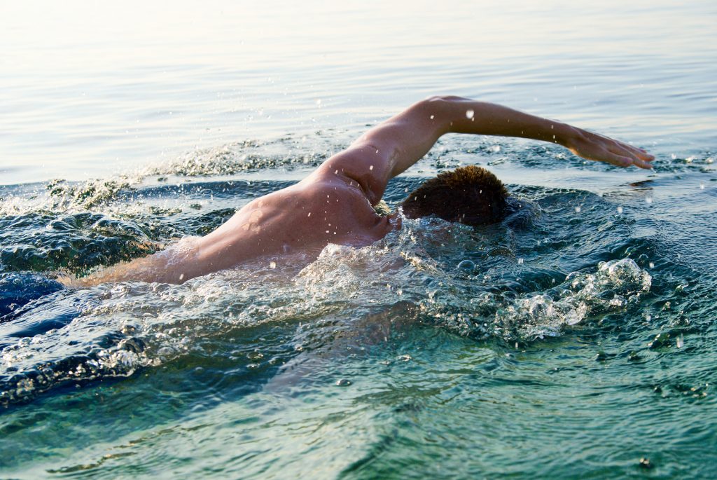 Person swimming freestyle in clear, sunlit water, their arm extended forward. Ripples and splashes surround, conveying movement and energy.