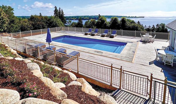 Scenic ocean view from a hillside deck featuring a rectangular pool, surrounded by lounge chairs and an umbrella, under a clear blue sky.