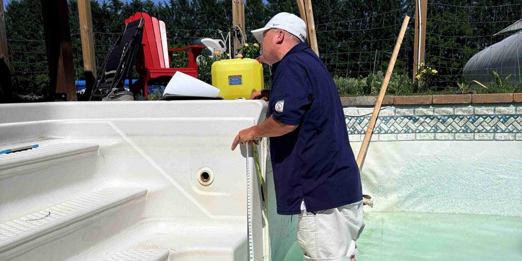 A man in a navy shirt and white cap stands in an empty pool using a measuring stick. Nearby are pool steps and a deck with chairs, suggesting maintenance.