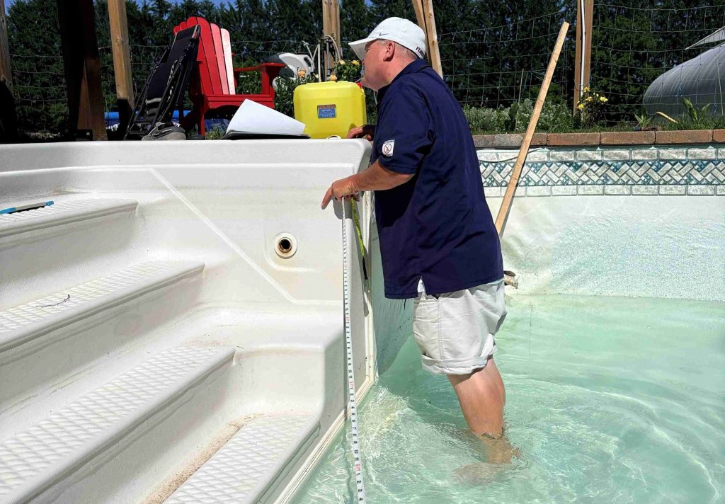 A man in a navy shirt and white cap stands in an empty pool using a measuring stick. Nearby are pool steps and a deck with chairs, suggesting maintenance.