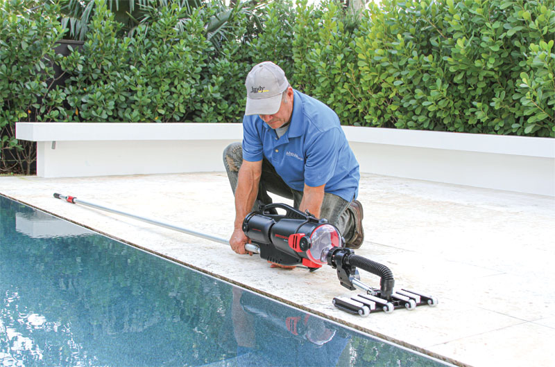 A man in a blue shirt and cap kneels by a pool, operating a pool cleaning device. Green shrubs surround the area, creating a calm atmosphere.
