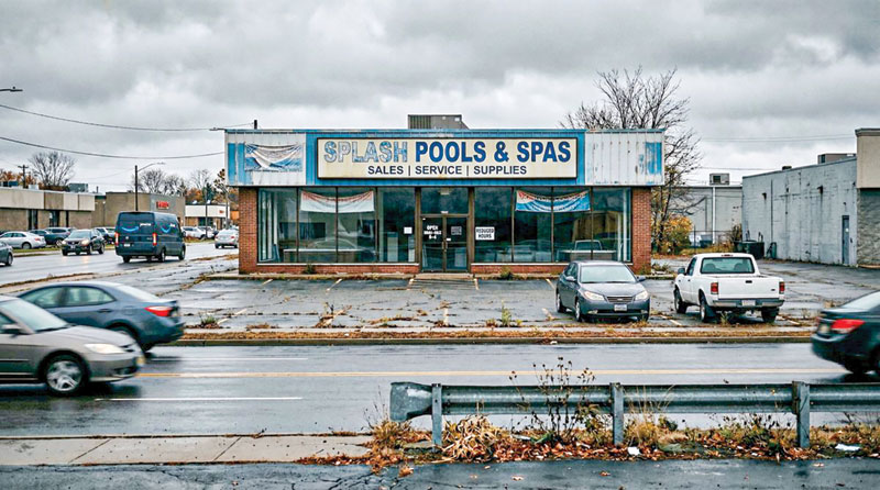 A closed "Splash Pools & Spas" store with a weathered sign sits in a drab, overcast urban area. Empty parking lot and moving cars convey a deserted tone.