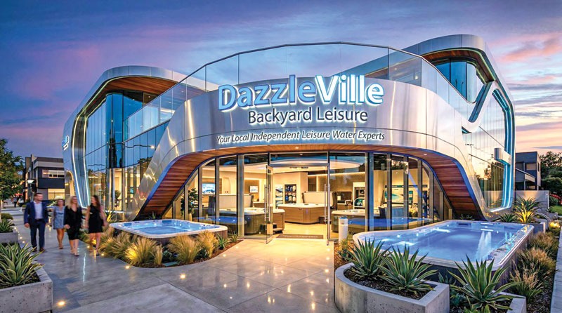 Modern showroom for "DazzleVille Backyard Leisure" at dusk, featuring sleek architecture, illuminated signage, and outdoor hot tubs. People walking nearby.
