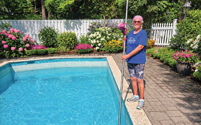 A person wearing a pink bandana and blue shirt smiles while cleaning a clear blue pool with a long pole. Vibrant flowers and green trees surround the pool area.
