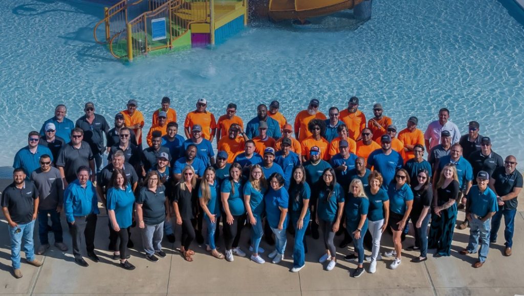 A group of diverse adults in blue and orange shirts stand smiling by a water park pool with slides in the background, conveying a cheerful atmosphere.