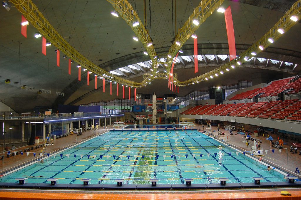 Spacious indoor swimming pool with lane dividers and diving platforms, surrounded by red seating. Bright overhead lights and red banners create a lively ambiance.