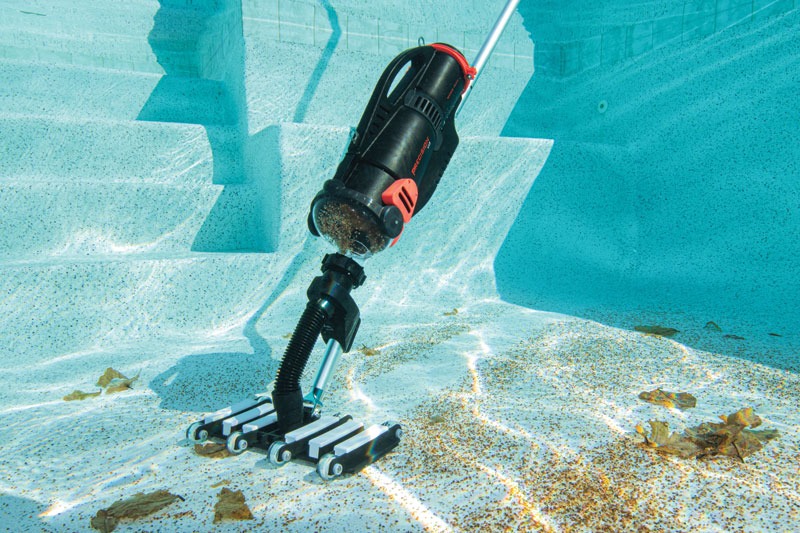 Underwater pool vacuum cleaning debris from a pool floor, surrounded by fallen leaves and dirt. Sunlight reflects on the blue pool steps.