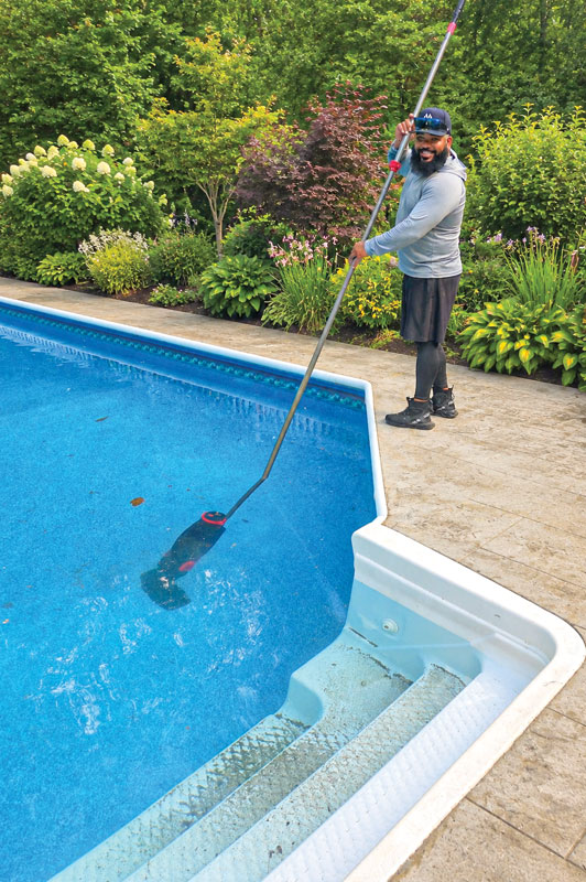 Man cleaning a bright blue pool with a long vacuum pole, surrounded by lush green and flowering bushes. The scene feels calm and refreshing.
