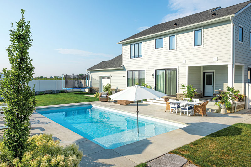 Two-story house with a beige facade and a dark roof, overlooking a sunny backyard with a blue pool. White patio furniture and an umbrella create an inviting, relaxing atmosphere.